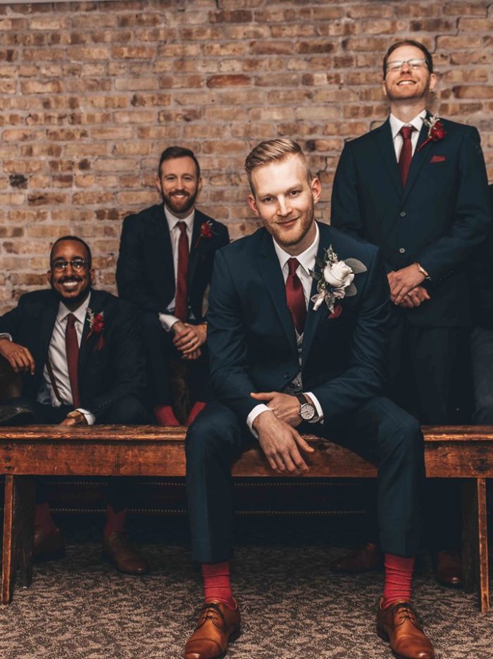 Groom sitting on wooden bench in front of groomsmen near brick wall